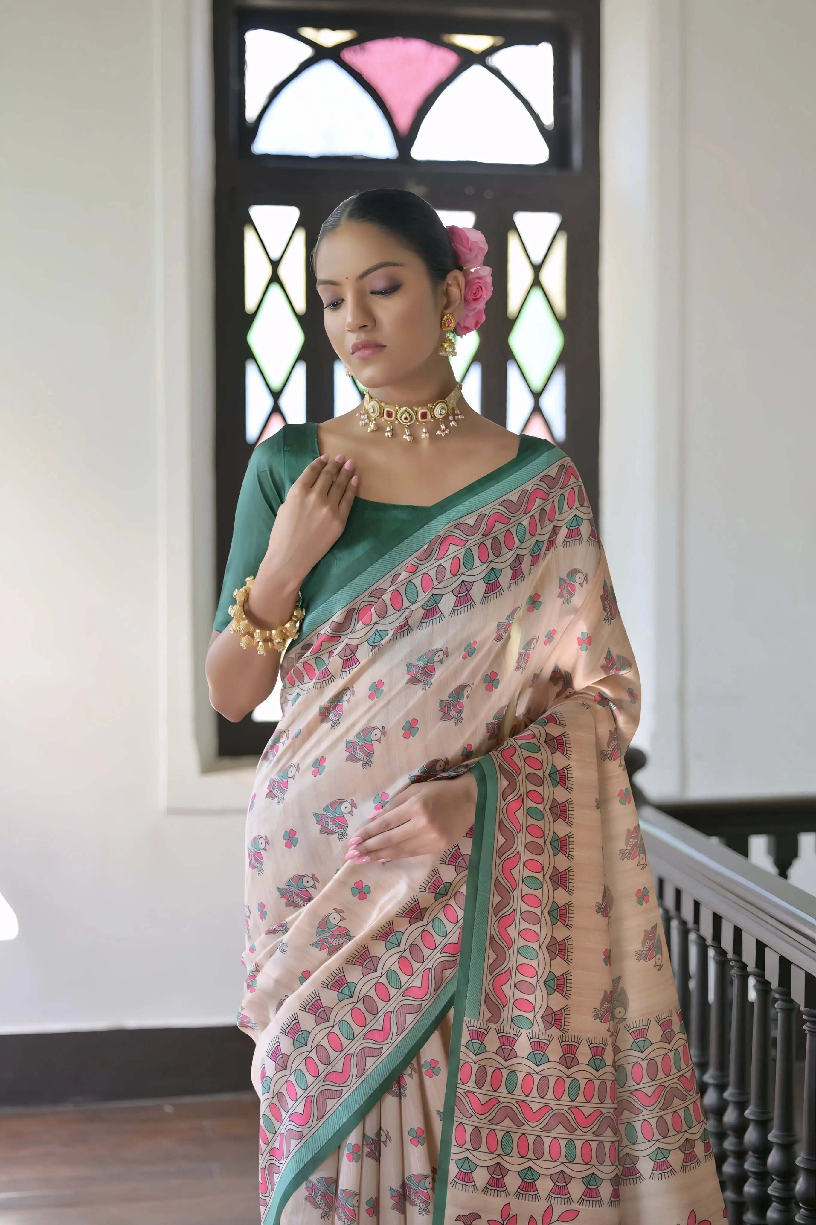 A woman models an elegant Off-White Soft Banarasi Silk Saree. The saree features a wide, prominent border and pallu with rich gold Zari weaving/print in shades of orange, peach, and green. She wears a coral sleeveless blouse and accessorizes with a gold Kundan choker necklace, large earrings, and a distinct dome-shaped handbag, posing indoors against a deep red backdrop.